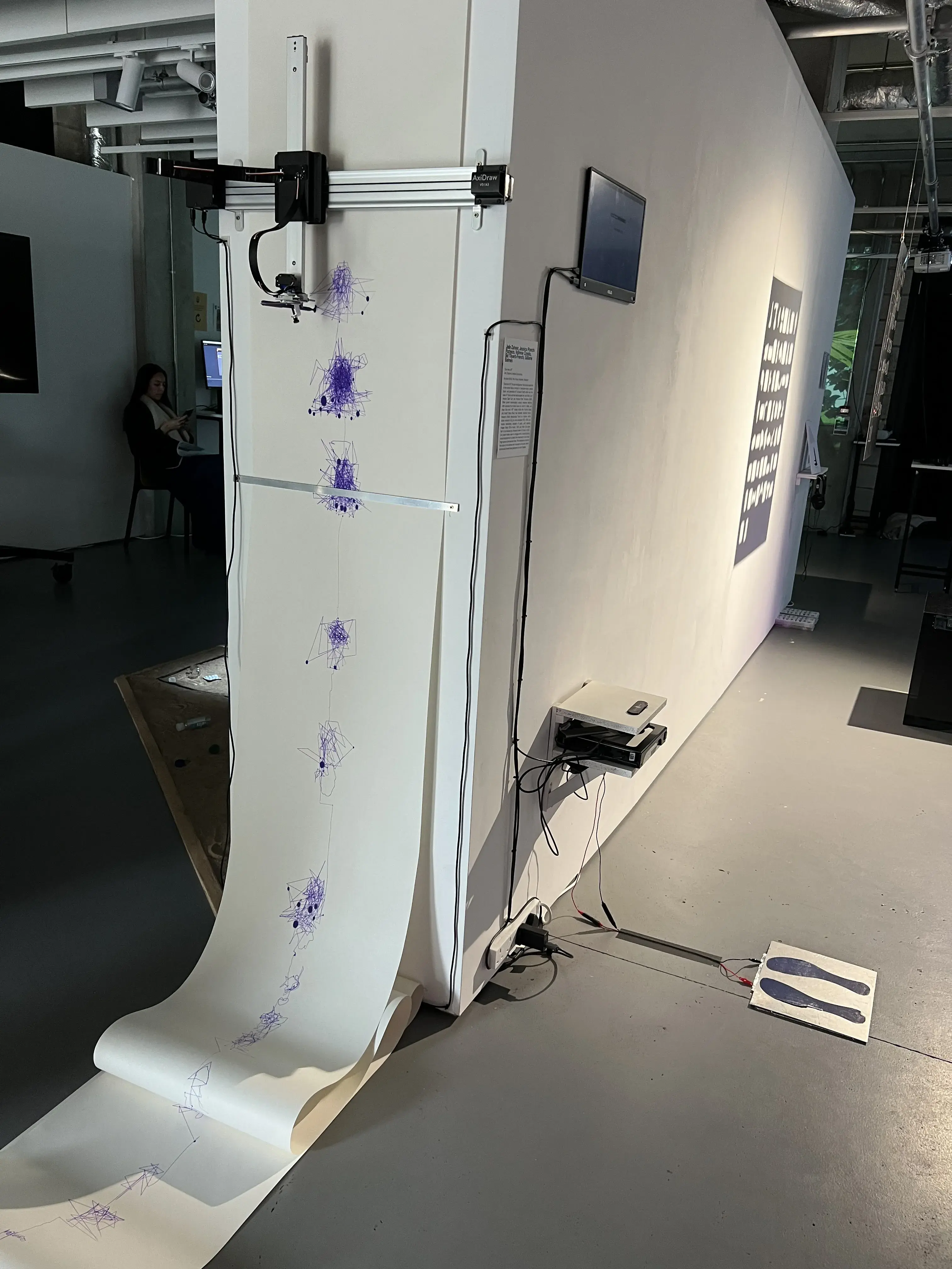 A pen plotter is mounted to a wall and a large roll of paper with drawings on it is coming out of it like a printer.